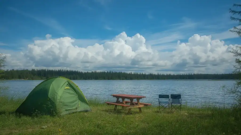 A green tent, a picnic table and two camp chairs on a grassy lakeshore under a blue sky and big white clouds